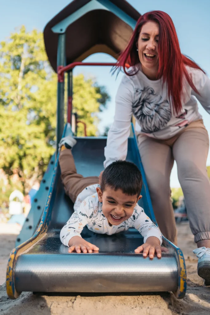 Child laughing while sliding down a playground slide headfirst, guided by an adult woman with red hair, on a sunny day at the park.