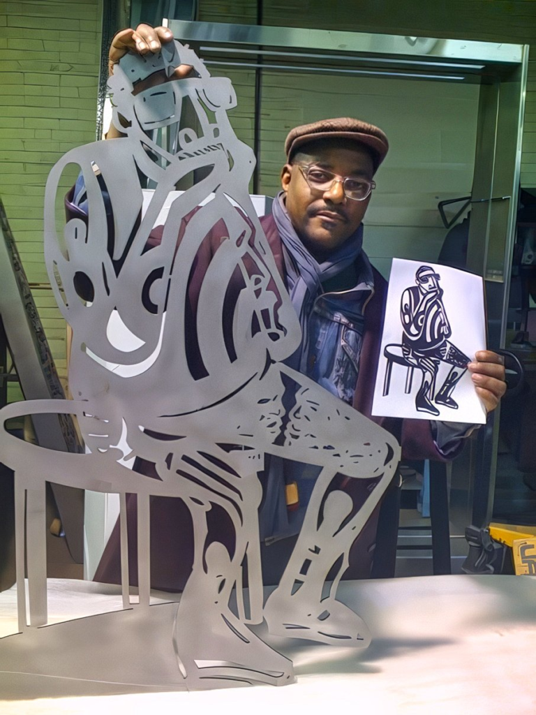 Artist Jerry Gant holding a printed sketch while standing beside a metal sculpture prototype in a workshop setting.