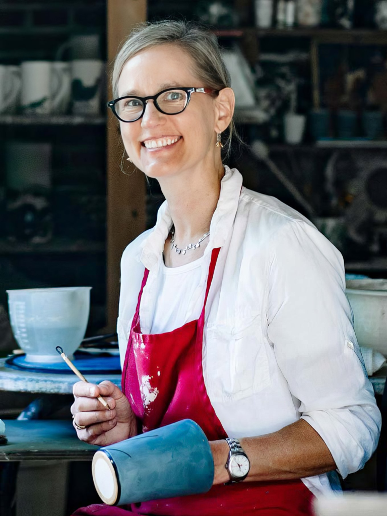 Katherine Hackl smiling in her ceramics studio, holding a hand-painted tile mug and brush, mosaic artist behind Nat Turner Park amphitheater installation